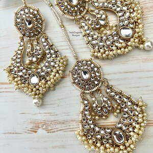 May include: A pair of gold-tone earrings with pearl accents. The earrings feature a large, round, mirrored center piece with intricate detailing. The earrings are suspended from a chain with a smaller, round, mirrored piece at the top. The earrings are accented with small, white pearls.