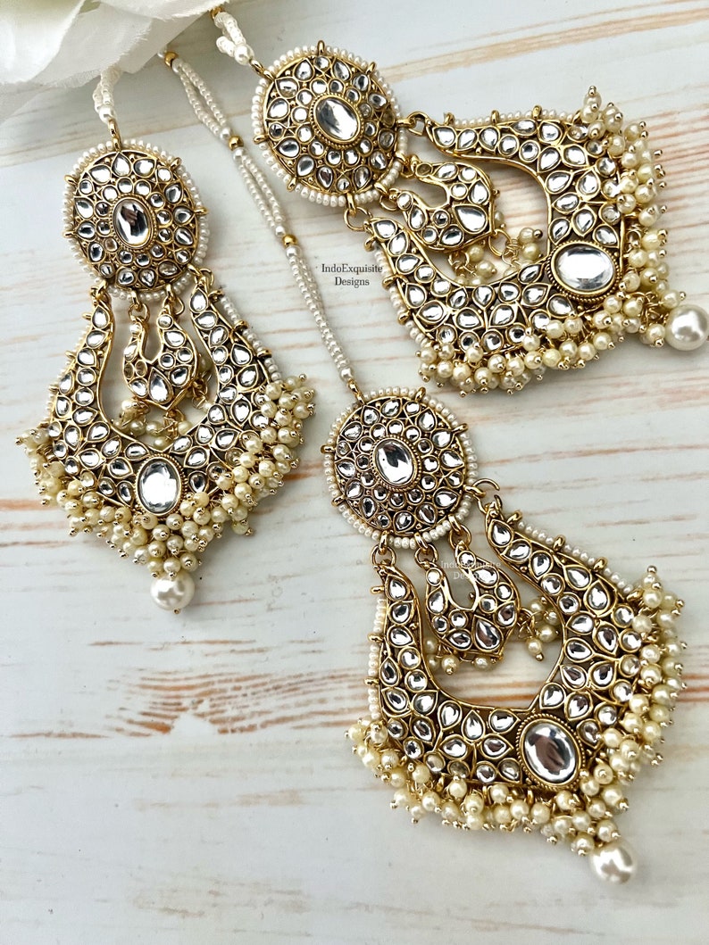 May include: A pair of gold-tone earrings with pearl accents. The earrings feature a large, round, mirrored center piece with intricate detailing. The earrings are suspended from a chain with a smaller, round, mirrored piece at the top. The earrings are accented with small, white pearls.