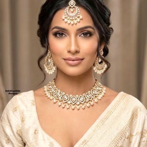 May include: A woman wearing a gold and pearl jewelry set, including a necklace, earrings, and a headpiece. The jewelry is set against a cream-colored outfit. The image is a close-up, showcasing the intricate details of the jewelry.