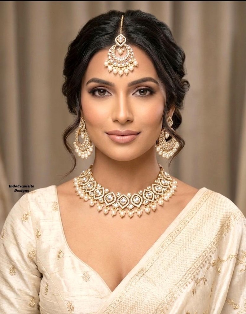 May include: A woman wearing a gold and pearl jewelry set, including a necklace, earrings, and a headpiece. The jewelry is set against a cream-colored outfit. The image is a close-up, showcasing the intricate details of the jewelry.