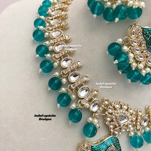May include: A teal and gold necklace and earrings set with a floral design and clear stones. The necklace has a large pendant with a square shape and the earrings are round. The set is made of metal and glass.