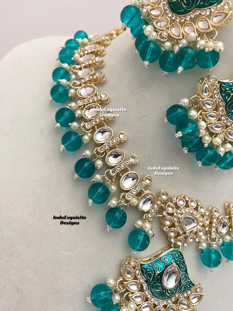 May include: A teal and gold necklace and earrings set with a floral design and clear stones. The necklace has a large pendant with a square shape and the earrings are round. The set is made of metal and glass.
