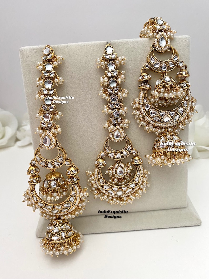 Puede incluir: Un conjunto de joyer&iacute;a en tono dorado que incluye dos pendientes largos y un adorno para la cabeza a juego, decorados con piedras claras reflectantes y peque&ntilde;as perlas blancas. Las piezas muestran metalister&iacute;a detallada.