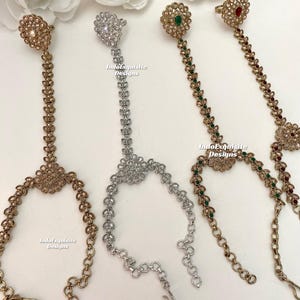 May include: Four gold and silver finger rings with dangling chains and decorative stones. The rings are adorned with clear and green stones. The chains are made of small linked circles. The text "IndoExquisite Designs" appears on the image.