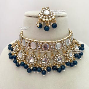 May include: A gold necklace with a large, round, mirrored pendant surrounded by smaller mirrored pendants. The necklace is adorned with blue beads and white pearls. The necklace is on a white display stand. IndoExquisite Designs is written on the stand.