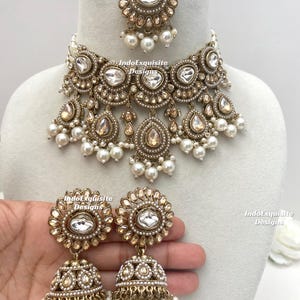 May include: An antique gold-toned jewelry set featuring a necklace, earrings, and a headpiece. The necklace and earrings have large, clear, oval-shaped stones and dangling teardrop-shaped stones. The earrings are held by a hand. The set is adorned with small pearls.