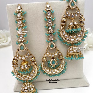 Puede incluir: Un conjunto de joyer&iacute;a dorado que incluye dos pendientes largos y un maang tikka central, adornados con piedras blancas, cuentas color verde azulado y colgantes jhumka en forma de campana. Las piezas llevan el texto IndoExquisite Designs.