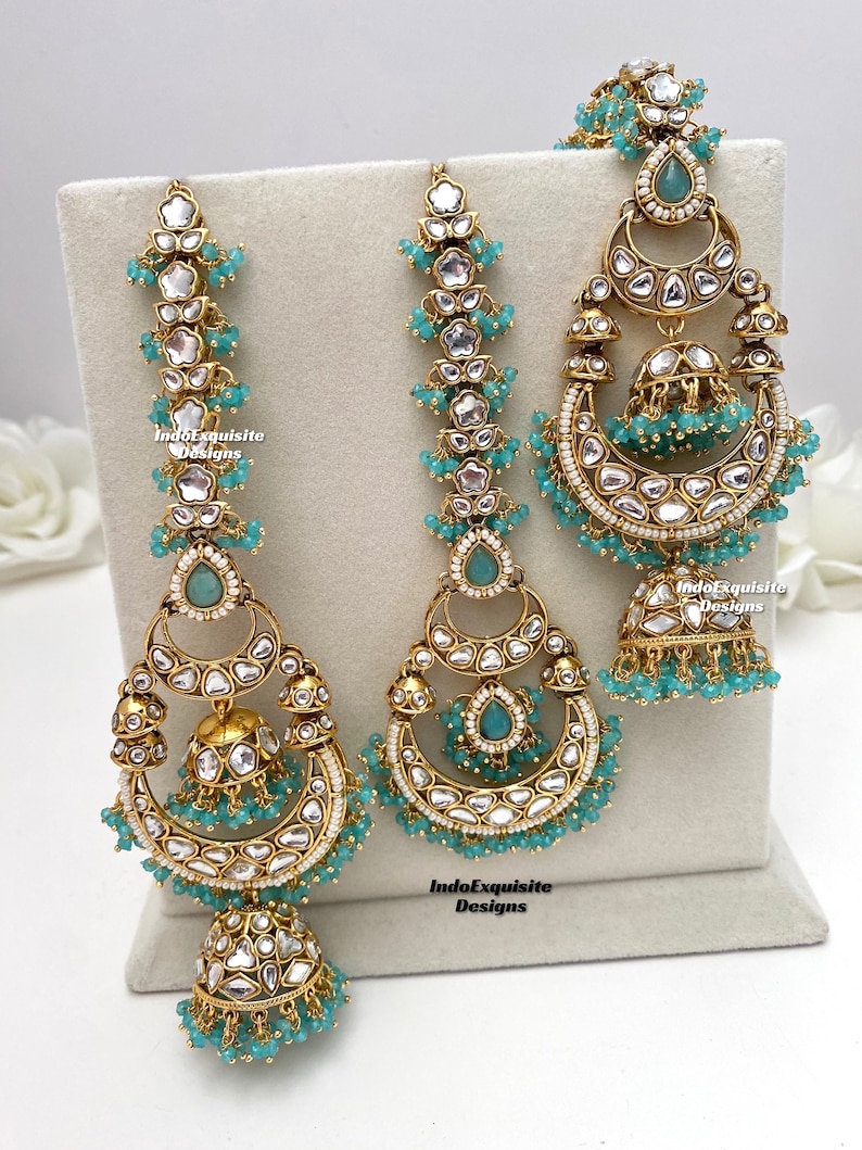 Puede incluir: Un conjunto de joyer&iacute;a dorado que incluye dos pendientes largos y un maang tikka central, adornados con piedras blancas, cuentas color verde azulado y colgantes jhumka en forma de campana. Las piezas llevan el texto IndoExquisite Designs.