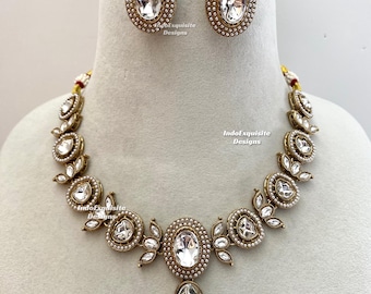 Puede incluir: Un conjunto de collar y pendientes en tono dorado con piedras ovaladas transparentes rodeadas de pequeñas perlas blancas. El collar tiene un colgante ovalado central con una piedra en forma de lágrima. El texto IndoExquisite Designs es visible.