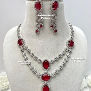 Puede incluir: Un deslumbrante conjunto de joyas con collar y pendientes a juego. El collar tiene dos capas con gemas rojas ovaladas y en forma de pera, rodeadas de pequeñas piedras transparentes. Los pendientes tienen un diseño similar, con gemas rojas ovaladas.