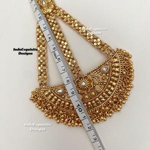 May include: A gold-colored headpiece with floral and beaded details. The headpiece is measured with a tape measure, showing a length of approximately 10 inches. The design includes a central floral motif and dangling beads.