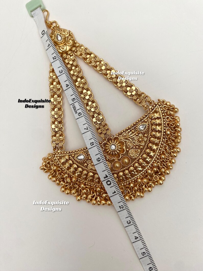 May include: A gold-colored headpiece with floral and beaded details. The headpiece is measured with a tape measure, showing a length of approximately 10 inches. The design includes a central floral motif and dangling beads.