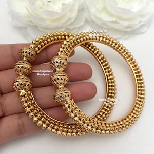 May include: Two gold bangles with detailed beaded patterns. The bangles have a textured surface with small, raised dots and spherical embellishments. The image includes the text "IndoExquisite Designs".