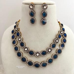 Puede incluir: Un collar y pendientes de dos niveles con piedras azules y blancas. El collar presenta una doble fila de piedras azules ovaladas rodeadas de piedras blancas más pequeñas. Los pendientes son pendientes colgantes con tres piedras azules.