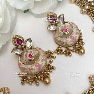 Pu&ograve; includere: Orecchini color oro con un design a mezzaluna color crema, adornati con motivi floreali rosa e una gemma rossa centrale. Gli orecchini presentano dettagli dorati pendenti e gemme trasparenti. Il testo "IndoExquisite Designs" &egrave; visibile.