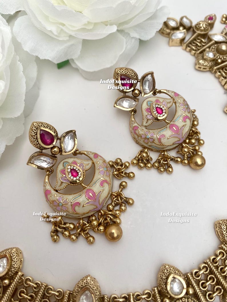 Pu&ograve; includere: Orecchini color oro con un design a mezzaluna color crema, adornati con motivi floreali rosa e una gemma rossa centrale. Gli orecchini presentano dettagli dorati pendenti e gemme trasparenti. Il testo "IndoExquisite Designs" &egrave; visibile.