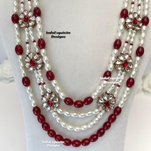 May include: A multi-strand necklace featuring white pearls and deep red beads. The necklace is accented with gold-toned floral pendants with red center stones. The text "IndoExquisite Designs" is visible.