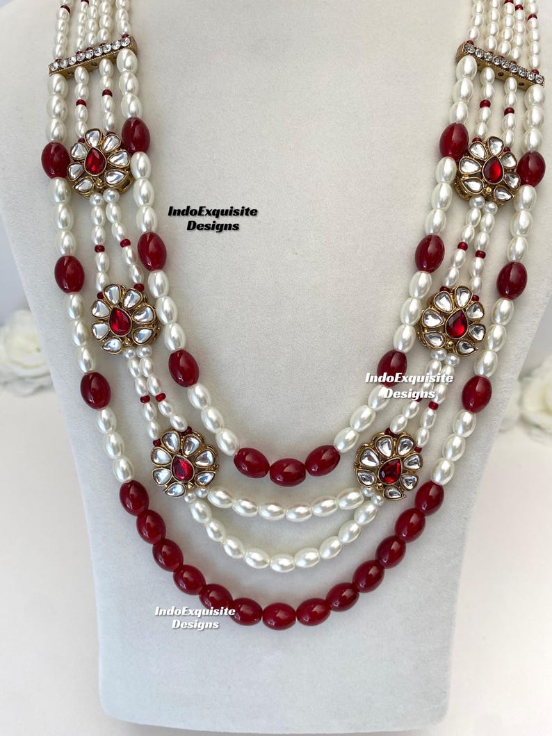 May include: A multi-strand necklace featuring white pearls and deep red beads. The necklace is accented with gold-toned floral pendants with red center stones. The text "IndoExquisite Designs" is visible.