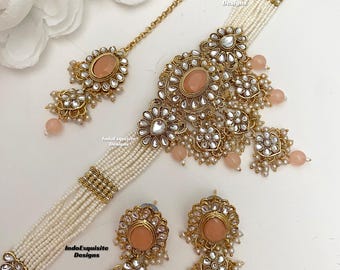 May include: A gold and white pearl choker necklace with a large, peach-colored stone pendant. The necklace is paired with matching earrings with peach-colored stones. The text "IndoExquisite Designs" is visible on the image.