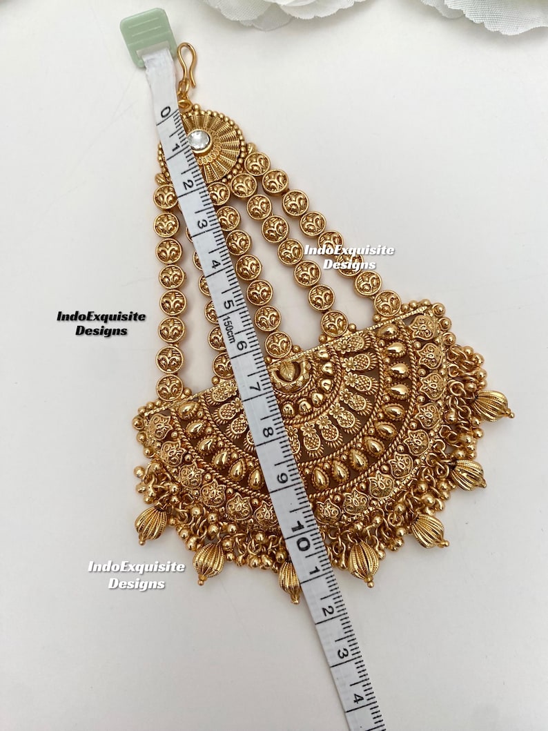 May include: A gold-toned headpiece featuring a central semi-circular pendant with intricate embossed patterns and dangling beads, attached to three chains with circular links. A measuring tape shows a length of approximately 4.5 inches.