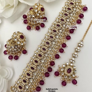 May include: A gold and white necklace with a large, ornate pendant and dangling pink beads. The necklace is paired with matching earrings and a matching maang tikka, a traditional Indian headpiece. The text "IndoExquisite Designs" appears three times on the image.