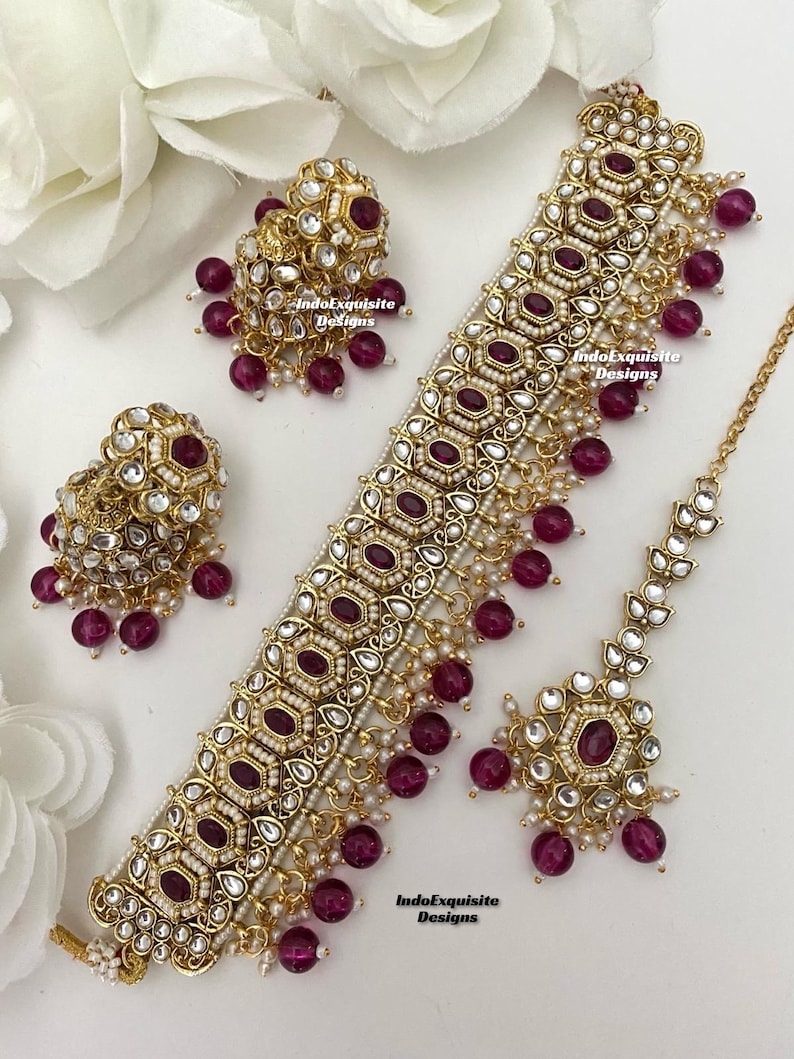 May include: A gold and white necklace with a large, ornate pendant and dangling pink beads. The necklace is paired with matching earrings and a matching maang tikka, a traditional Indian headpiece. The text "IndoExquisite Designs" appears three times on the image.