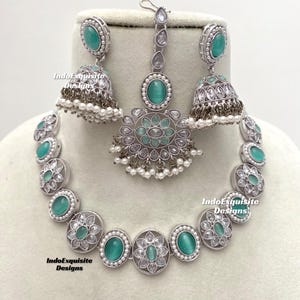 Op de afbeelding: Een zilveren ketting met een hanger en bijpassende oorbellen. De ketting en oorbellen hebben ronde, turquoise stenen omgeven door kleinere, heldere stenen. De hanger heeft een druppelvorm met een vergelijkbaar ontwerp.