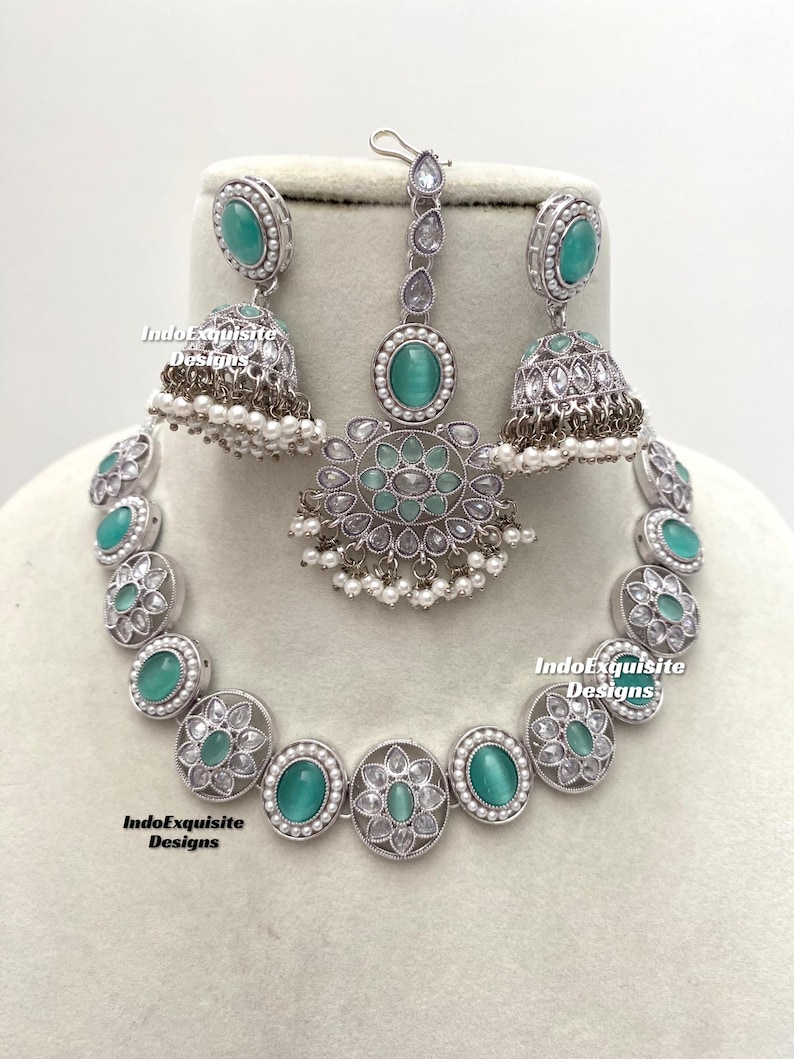 Op de afbeelding: Een zilveren ketting met een hanger en bijpassende oorbellen. De ketting en oorbellen hebben ronde, turquoise stenen omgeven door kleinere, heldere stenen. De hanger heeft een druppelvorm met een vergelijkbaar ontwerp.