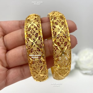 May include: Two gold bangles with detailed floral and geometric cut-out patterns. The bangles are thick and have a shiny finish. The brand name "IndoExquisite Designs" is visible on the bangles.