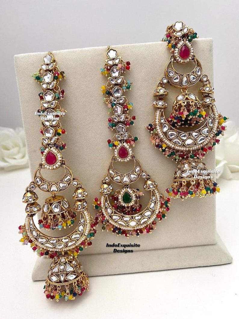 Puede incluir: Tres pendientes dorados con intrincados trabajos en metal, piedras transparentes y cuentas de colores. Los dise&ntilde;os incluyen formas de media luna, elementos en forma de campana y piedras centrales en forma de l&aacute;grima en rosa y verde, marcados IndoExquisite Designs.
