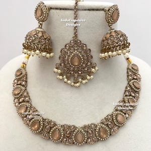 May include: A complete jewelry set featuring a necklace, earrings, and a headpiece. The set has a vintage aesthetic with a bronze-colored metal and peach-colored stones. The earrings are bell-shaped with dangling pearls. The necklace and headpiece have a similar design.