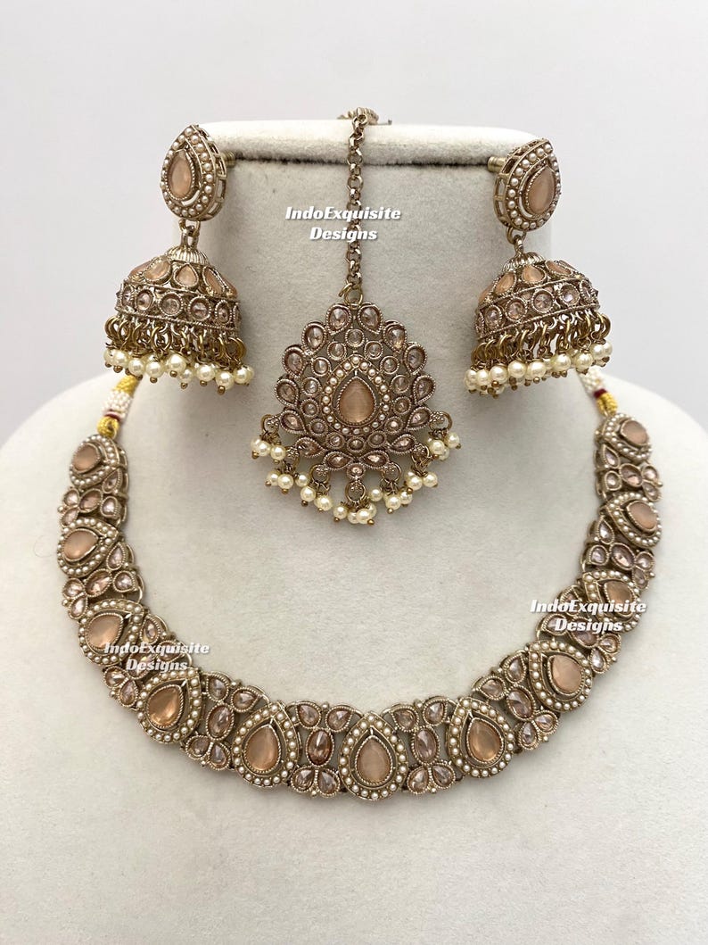 May include: A complete jewelry set featuring a necklace, earrings, and a headpiece. The set has a vintage aesthetic with a bronze-colored metal and peach-colored stones. The earrings are bell-shaped with dangling pearls. The necklace and headpiece have a similar design.