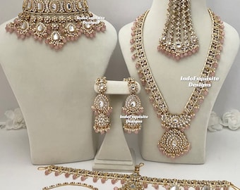 May include: A bridal jewelry set with a gold tone finish and pink accents. The set includes a necklace, earrings, a maang tikka, a choker, a nose ring, and a bracelet.