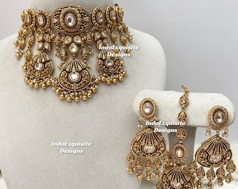 Puede incluir: Un conjunto de joyería en tono dorado antiguo que incluye un collar tipo gargantilla, dos pendientes colgantes y un maang tikka. Cada pieza tiene grabados florales, piedras blancas y pequeñas cuentas colgantes. El texto dice IndoExquisite Designs.