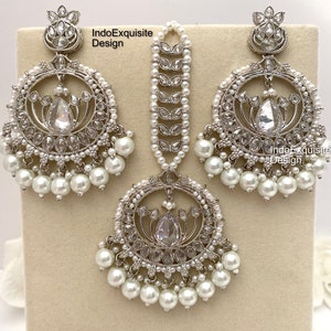 May include: A silver-toned jewelry set featuring a pair of large, round earrings and a matching headpiece. Each piece is adorned with clear crystals and faux pearls. The earrings and headpiece have a detailed, ornate design, with the text "IndoExquisite Design" visible.