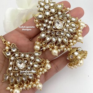 May include: Gold-toned jewelry set featuring intricate floral and geometric designs. The set includes a headpiece and earrings, adorned with clear crystals and faux pearls. The jewelry is displayed on a light background.