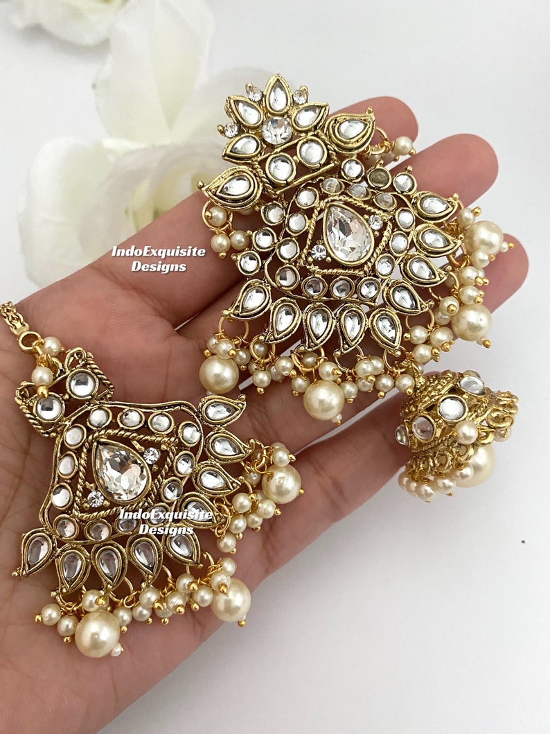 May include: Gold-toned jewelry set featuring intricate floral and geometric designs. The set includes a headpiece and earrings, adorned with clear crystals and faux pearls. The jewelry is displayed on a light background.
