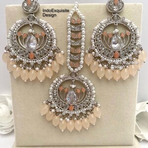 May include: An ornate, three-piece jewelry set featuring a silver-toned metal design. The set includes a pair of large hoop earrings, a headpiece, and a pendant. Each piece is adorned with clear and peach-colored stones, small pearls, and peach-colored teardrop beads. The text "IndoExquisite Design" is visible.