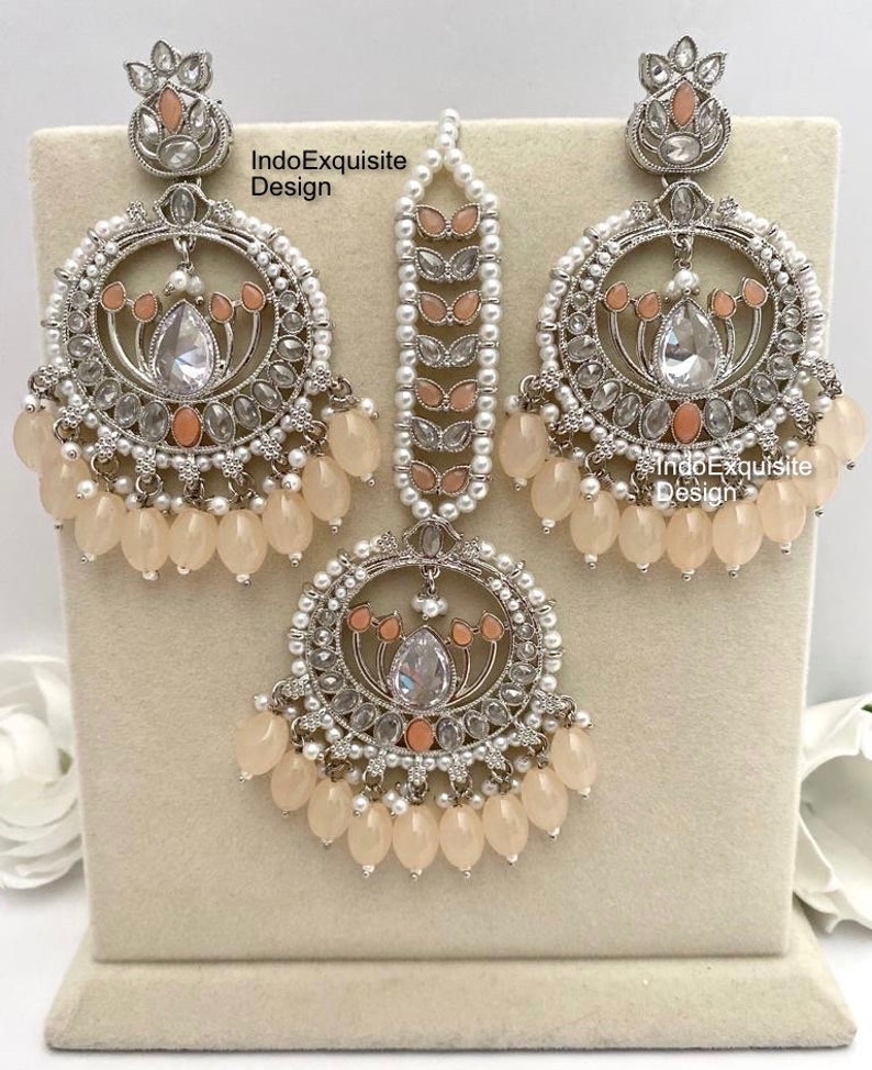 May include: An ornate, three-piece jewelry set featuring a silver-toned metal design. The set includes a pair of large hoop earrings, a headpiece, and a pendant. Each piece is adorned with clear and peach-colored stones, small pearls, and peach-colored teardrop beads. The text "IndoExquisite Design" is visible.