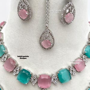 May include: A silver necklace, earrings, and a pendant with turquoise and pink stones. The stones are surrounded by clear rhinestones. The jewelry is set on a white background. IndoExquisite Designs is written on the background.