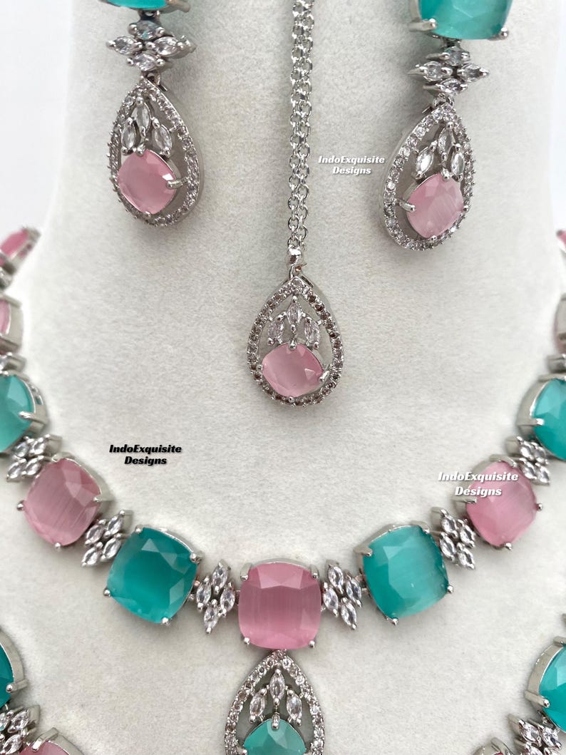 May include: A silver necklace, earrings, and a pendant with turquoise and pink stones. The stones are surrounded by clear rhinestones. The jewelry is set on a white background. IndoExquisite Designs is written on the background.