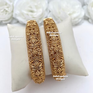 May include: Two gold bangles with detailed floral and leaf designs. The bangles are presented on a white cushion, with white flowers in the background. The text "IndoExquisite Designs" is visible on the cushion.