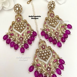 Puede incluir: Un juego de pendientes de tono dorado con un diseño de esmalte blanco y rosa, con forma de corazón y cuentas moradas colgantes. Los pendientes se combinan con un collar a juego.