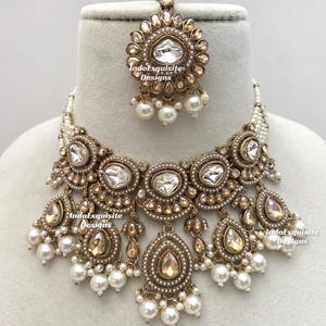 May include: An ornate gold-toned necklace and headpiece set. The necklace features large, clear, and teardrop-shaped stones, surrounded by smaller stones and pearls. The headpiece mirrors the necklace design. The set is displayed on a white surface.