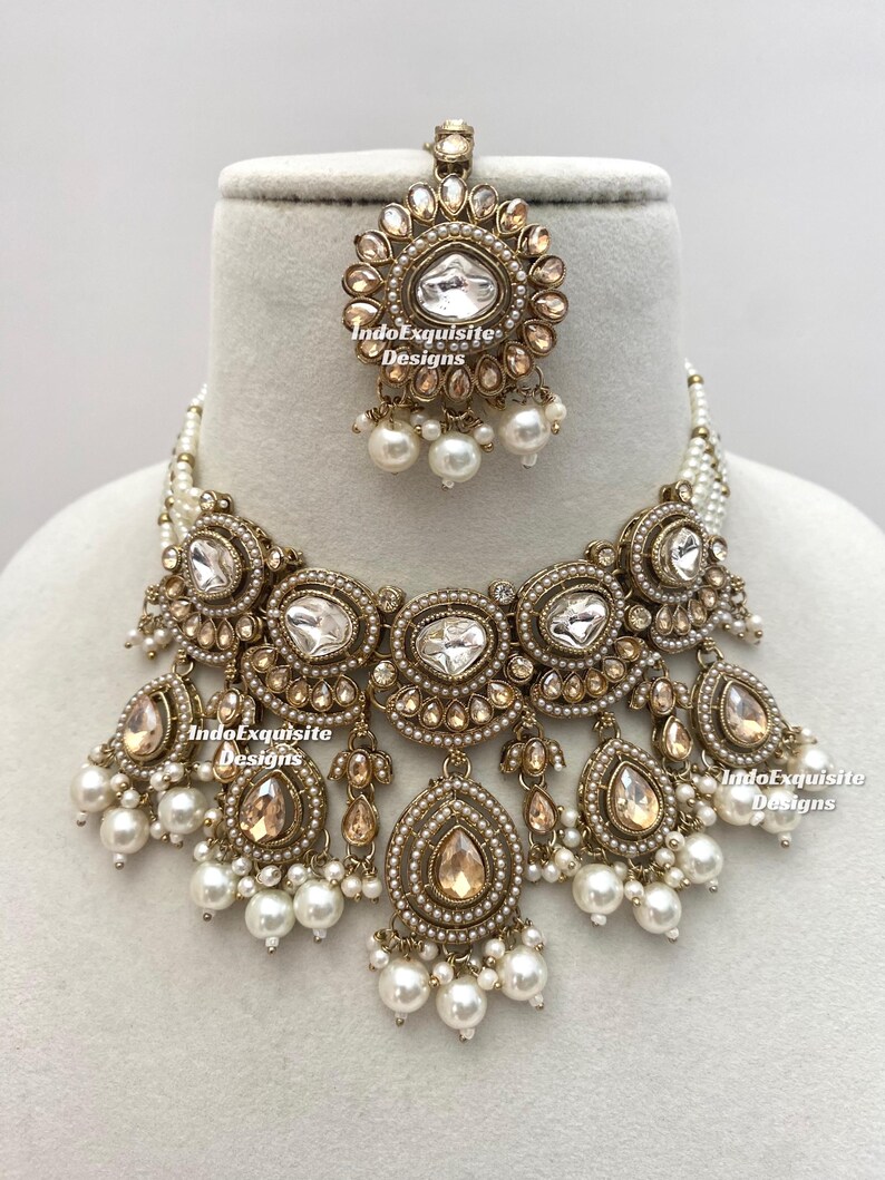 May include: An ornate gold-toned necklace and headpiece set. The necklace features large, clear, and teardrop-shaped stones, surrounded by smaller stones and pearls. The headpiece mirrors the necklace design. The set is displayed on a white surface.