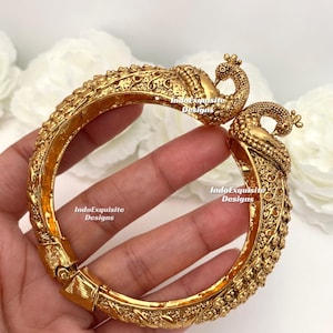 May include: Gold-colored bangle bracelet with intricate embossed detailing. The bracelet features a peacock design at the clasp, with the peacock's head and tail feathers forming the closure. The bracelet is held in a hand.