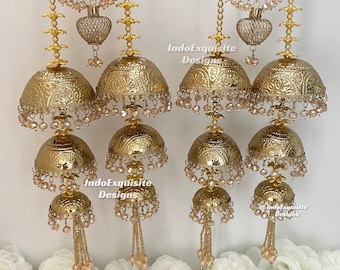 Puede incluir: Un par de adornos colgantes dorados con múltiples dijes jhumka en forma de campana escalonada con patrones en relieve, acentuados por cuentas claras colgantes y pequeñas esferas de malla metálica, marcados IndoExquisite Designs.