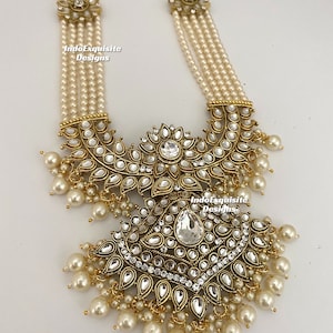 May include: An ornate necklace featuring multiple strands of faux pearls. The necklace has a large, detailed pendant with clear and white gemstones set in a gold-toned metal. The pendant is adorned with dangling pearl accents. The text "IndoExquisite Designs" is visible.