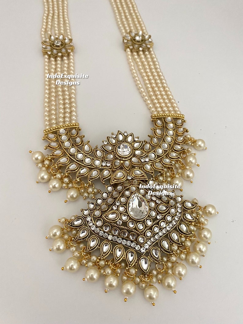 May include: An ornate necklace featuring multiple strands of faux pearls. The necklace has a large, detailed pendant with clear and white gemstones set in a gold-toned metal. The pendant is adorned with dangling pearl accents. The text "IndoExquisite Designs" is visible.