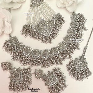 May include: A silver-toned bridal jewelry set featuring a necklace, earrings, tikka, and a headpiece. Each piece is adorned with small, round, white pearls and intricate filigree designs. The set includes the text "IndoExquisite Designs".
